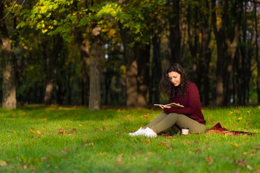 A cute girl reads a book and drinks coffee on a green lawn in an autumn park. Autumn mood. A cozy place to be alone with yourself.