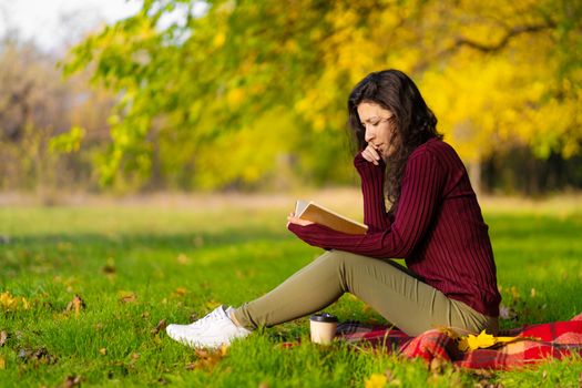 A cute girl reads a book and drinks coffee on a green lawn in an autumn park. Autumn mood. A cozy place to be alone with yourself.