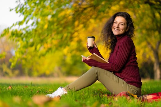 A cute girl reads a book and drinks coffee on a green lawn in an autumn park. Autumn mood. A cozy place to be alone with yourself.