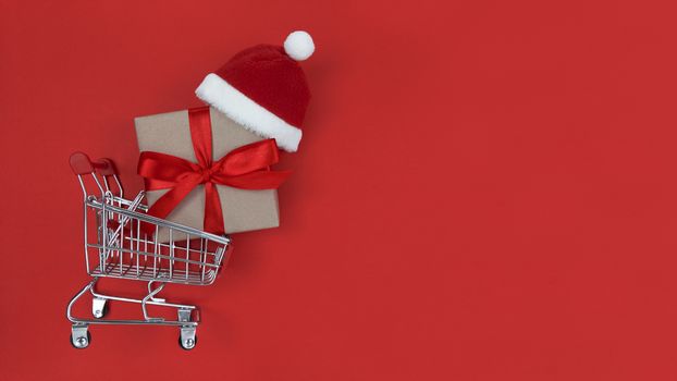 Supermarket trolley, gift box and Santa hat on red background.