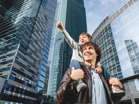 Little boy sits on father's shoulders and points on skyscraper. Dad and son looks on glass walls of buildings. Future and modern technologies, life balance and family life in well keeps districts.