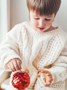 Kid with red decorative ball for Christmas tree. Boy in cable-knit oversized sweater. Cozy outfit for snuggle weather. Winter holiday spirit. New year.