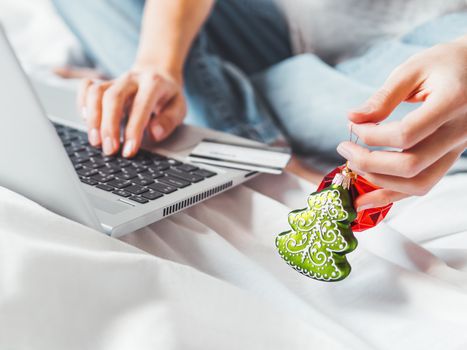 Woman using laptop to pay for online order by credit card. Decorative Christmas tree and New Year red ball in her hand. Online shopping before winter holidays.