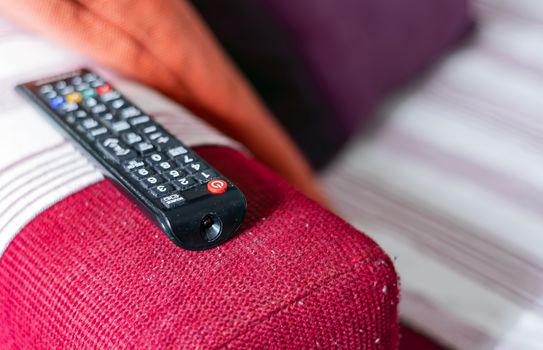 the dusty television remote control on the armrest of a red fabric sofa. Lint on the sofa. Sedentary life and home entertainment. Selective focus on the remote control