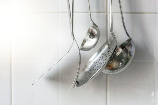 A skimmer, two forks and two metal ladles hanging on the tiled kitchen wall. Metal kitchen utensils