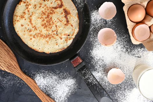 Hot pancake in black pan on black table with flour, milk and eggs.