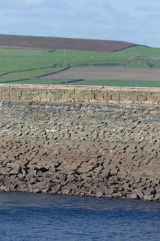 Vertical image of stone dam reservoir wall. Deep blue water and rural agricultural background are selectively out of focus. Warm sunny summers day