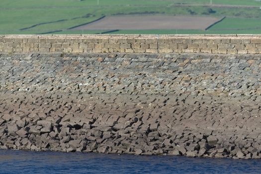 A rural large stone dam acts as the wall to a deep blue water reservoir. Selective focus blurs the agricultural fields in the background. Warm sun.
