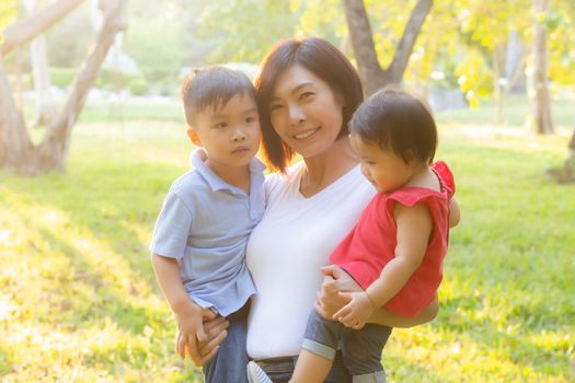 Beautiful young asian mother carrying little boy and girl in the park, asia woman happy having son and daughter and hug kid, mom love and embracing child together, mother day and family concept.