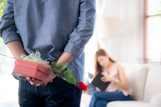 Beautiful young asian couple with man hiding red gift box and rose flower for surprise at home, husband and wife with happy valentine or birthday, married together, family love concept.