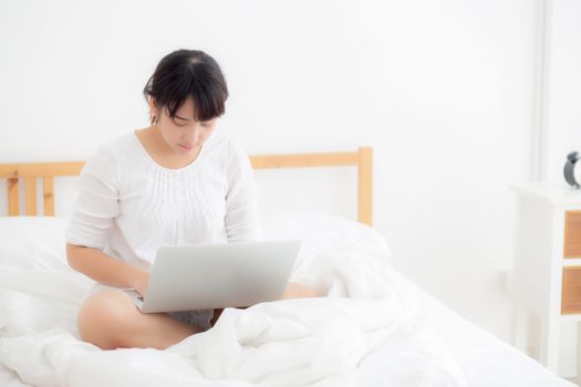 Portrait of beautiful asian young woman sitting on bed using laptop computer at bedroom for leisure and relax, freelance with girl working notebook, communication and lifestyle concept.