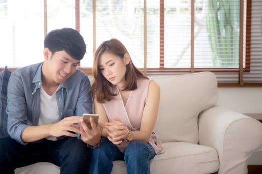 Young asian couple or friend looking smart mobile phone with enjoy at living room, happy family relax and fun media entertainment on smartphone on sofa together at home, lifestyle concept.