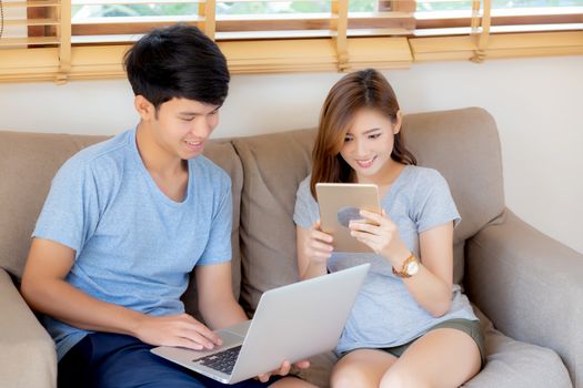 Beautiful young asian couple cheerful freelance working with man using laptop and woman using tablet on couch, family relax and sharing communication together at home, lifestyle concept.