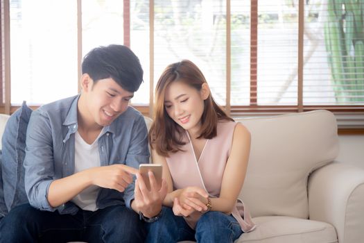 Young asian couple or friend looking smart mobile phone with enjoy at living room, happy family relax and fun media entertainment on smartphone on sofa together at home, lifestyle concept.