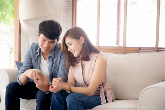 Young asian couple or friend looking smart mobile phone with enjoy at living room, happy family relax and fun media entertainment on smartphone on sofa together at home, lifestyle concept.