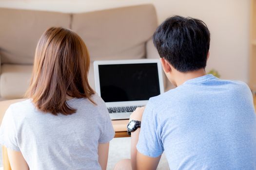 Back view of young asian couple working laptop with blank screen display, family planning and searching content together, man and woman looking computer, business and communication concept.