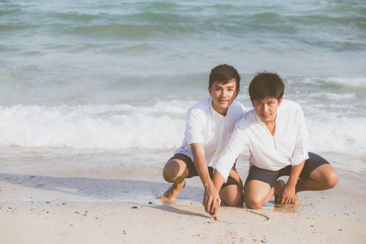 Young asian couple gay smiling romantic drawing heart shape together on sand in vacation, homosexual happy and fun with love sitting on sand at the beach in travel summer, LGBT legal concept.