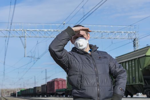 A foreman in a medical mask at a railway station looks out for trains, covering his face with his hand from the sun