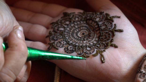 Close up macro shot of mehendi henna tattoo being applied in an intricate pattern on the hand of a woman with soft natural light and on a red background on the event of karwachauth marriage shaadi. Shows the very popular temporary tattoo method that is a part of the hindu religion and is widely done across india