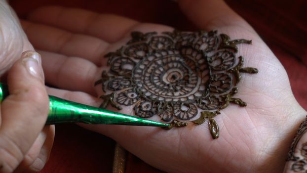 Close up macro shot of mehendi henna tattoo being applied in an intricate pattern on the hand of a woman with soft natural light and on a red background on the event of karwachauth marriage shaadi. Shows the very popular temporary tattoo method that is a part of the hindu religion and is widely done across india