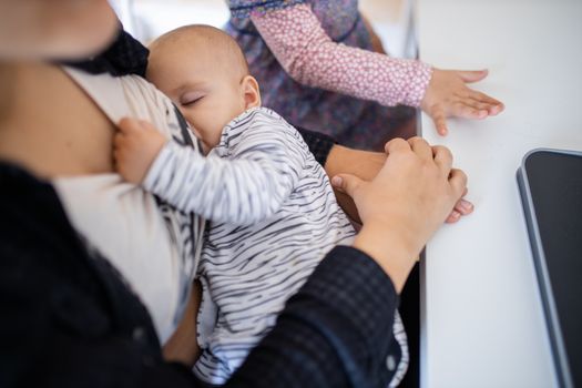 Sleeping baby peacefully resting in arms and holding the shirt of her mother who sits at a desk. Mother holding her slipping baby while using a laptop. Peacefully sleeping babies in arms