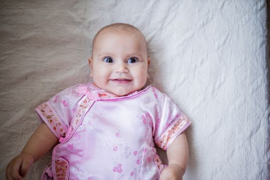 Happy smiling baby girl from above in Asian pink attire lying down on white bed. Portrait of joyful female baby resting on bed. Happy babies on cradles and beds