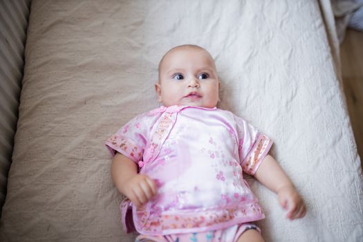 Curious adorable baby girl from above in Asian pink attire lying down on white bed. Portrait of cute female baby resting on bed. Happy babies on cradles and beds