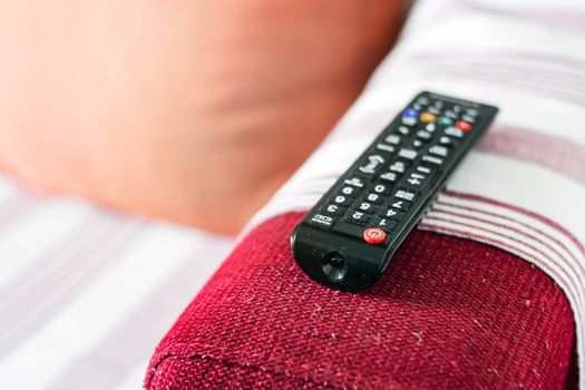 the dusty television remote control on the armrest of a red fabric sofa. Lint on the sofa. Sedentary life and home entertainment. Selective focus on the remote control