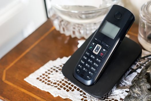 a black plastic cordless phone charging on its cradle inside a classically furnished home with antiques and white crochet. Outdated technology