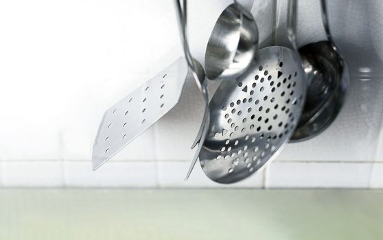 A skimmer, two forks and two metal ladles hanging on the tiled kitchen wall. Metal kitchen utensils