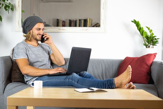 Handsome young business caucasian man working at home using laptop and talking with mobile phone