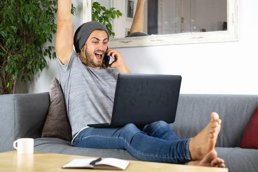 Handsome young business caucasian man working at home using laptop and talking with mobile phone feeling happy