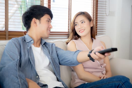 Beautiful young asian couple holding remote and watching tv or video streaming on sofa with relax and happy in living room at home, lifestyle family leisure with entertainment enjoy together on couch.