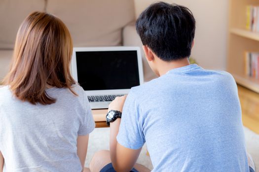 Back view of young asian couple working laptop with blank screen display, family planning and searching content together, man and woman looking computer, business and communication concept.