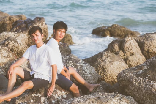 Homosexual portrait young asian couple sitting hug together on rock or stone in the beach in summer, asia gay going tourism for leisure and relax with happy in vacation at sea, LGBT legal concept.