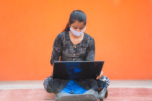 Young girl student in medical mask busy working on laptop at caollege corridor or universtiy campus - concept of college reopen, new normal due to coronavirus or covid-19 pandemic