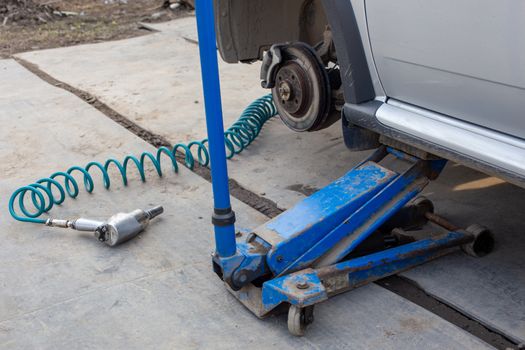 Car without wheel lifted with hydraulic floor jack and pneumatic impact wrench in a outdoors car workshop. Season tire changing procedure. Close view with selective focus and background blur.