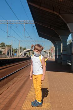 Little girl in a white T-shirt and jeans and a medical mask waiting for the train on the railway station. Keep social distancing to avoid the spread of COVID-19. Safe travel.