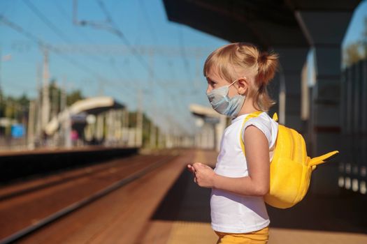 Little girl in a white T-shirt and jeans and a medical mask waiting for the train on the railway station. Keep social distancing to avoid the spread of COVID-19. Safe travel.