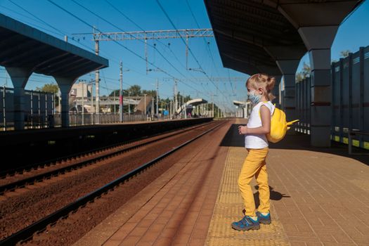 Little girl in a white T-shirt and jeans and a medical mask waiting for the train on the railway station. Keep social distancing to avoid the spread of COVID-19. Safe travel.