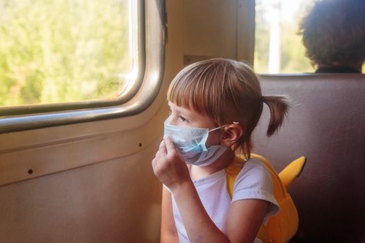 Little girl in a white T-shirt and jeans and a medical mask is sitting in the train. Keep social distancing to avoid the spread of COVID-19. Safe travel.