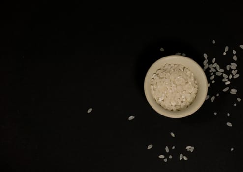 White uncooked rice in a beige bowl with scattered grains nearby on a black background