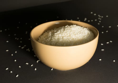 White uncooked rice in a beige bowl with scattered grains nearby on a black background