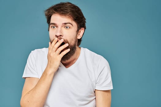 A bearded man in a white T-shirt gestures with his hands emotions blue background. High quality photo