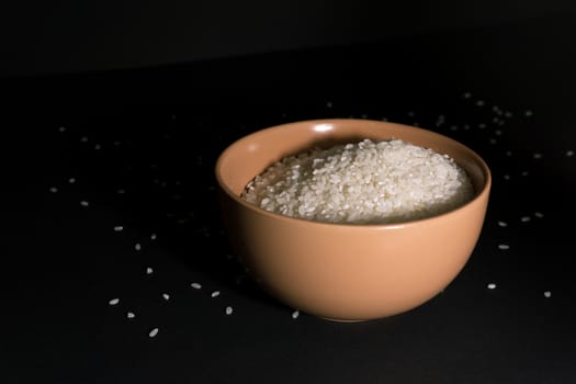 White uncooked rice in a beige bowl with scattered grains nearby on a black background