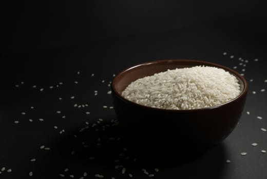 White uncooked rice in a brown bowl with scattered grains nearby on a black background