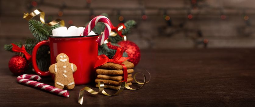 Cocoa in red mug with marshmallows candy cane fir tree branches and red baubles on dark wooden background with bokeh lights