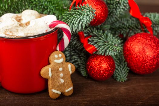Cocoa hot chocolate in red mug with marshmallows candy cane gingerbread cookie man fir tree branches and red baubles on dark wooden background