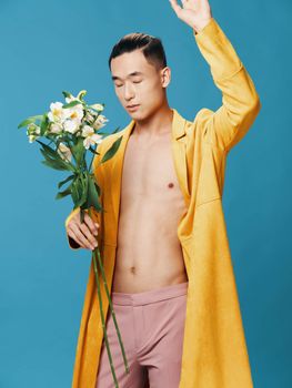 Young man of Asian appearance in a yellow coat with a bouquet of flowers gesturing with his hands on a blue background cropped view. High quality photo