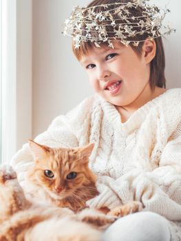 Kid with decorative star tinsel for Christmas tree hugs cute ginger cat. Boy in cable knit oversized sweater with fluffy pet. Winter holiday spirit. New year.